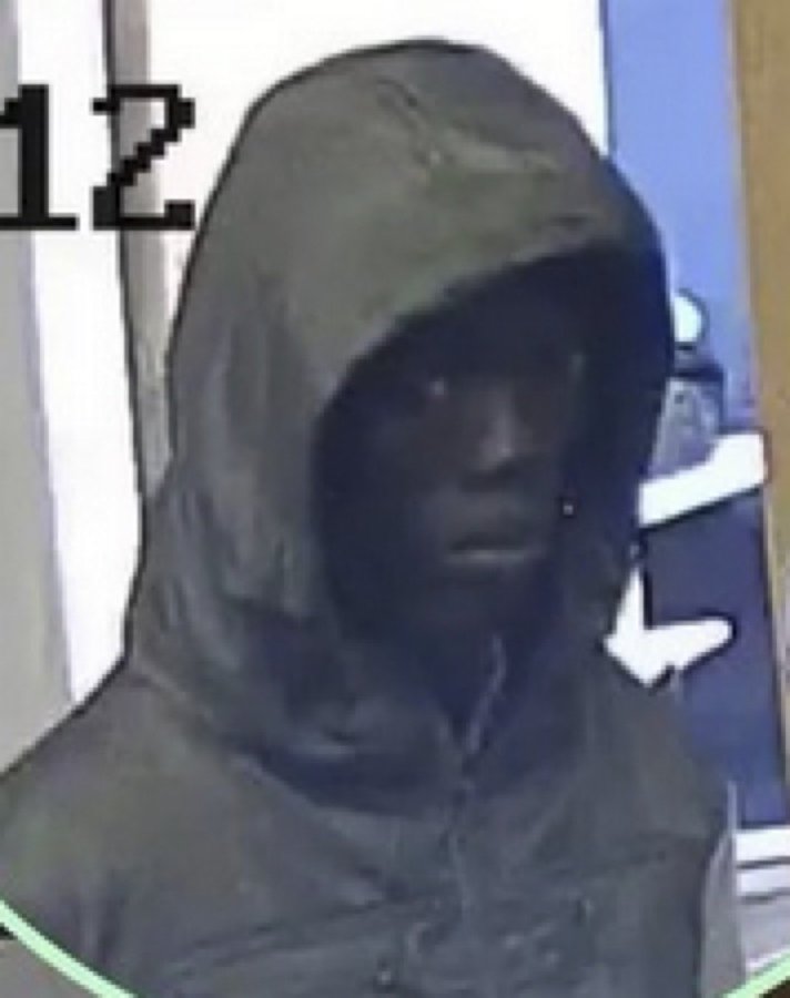 Image shows a person wearing a dark hooded sweatshirt with the hood up obscuring part of the face in a green-tinted surveillance style from a low angle in what appears to be a parking lot or outdoor area with a blue building and white markings on the ground.