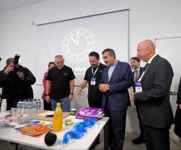 First image shows a group of men and women in professional attire standing around a table with water bottles, notebooks, and colorful items like yellow bottles and blue strings in a room with a projected clock on a screen and name badges. Second image depicts a diverse group of people including men and women in suits and casual clothes gathered in a modern room with white walls, chairs, and tables, some shaking hands and interacting. Third image features a bald man in a suit speaking near a whiteboard in a spacious room with wooden ceiling, surrounded by attendees in various outfits sitting at tables with cups and notebooks. Fourth image displays a group of about ten professionals in formal wear posing in front of a building entrance with a banner reading Zekâ ve Yetenek Atölyeleri and a modern architectural facade.