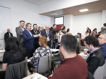 First image shows a group of men and women in professional attire standing around a table with water bottles, notebooks, and colorful items like yellow bottles and blue strings in a room with a projected clock on a screen and name badges. Second image depicts a diverse group of people including men and women in suits and casual clothes gathered in a modern room with white walls, chairs, and tables, some shaking hands and interacting. Third image features a bald man in a suit speaking near a whiteboard in a spacious room with wooden ceiling, surrounded by attendees in various outfits sitting at tables with cups and notebooks. Fourth image displays a group of about ten professionals in formal wear posing in front of a building entrance with a banner reading Zekâ ve Yetenek Atölyeleri and a modern architectural facade.