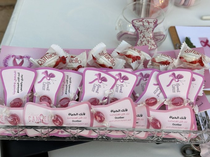 First image shows a group of women in traditional attire standing together outdoors near beige buildings with the charity logo and pink ribbon symbol for breast cancer awareness overlaid on a promotional banner reading awareness campaign for breast cancer early detection. Second image displays a table with pink-themed items including wrapped sweets labeled sweet love, cups with pink ribbon designs, and stacks of promotional materials for breast cancer awareness. Third image depicts an outdoor setup against a traditional building with a large pink ribbon standee, vases of flowers, a table with pink cups, jars of drinks, and sign-up sheets for the awareness event. Fourth image features a woven basket on a table filled with small gift bags containing perfume bottles and pink ribbon tags labeled gifts for breast cancer awareness campaign participants.