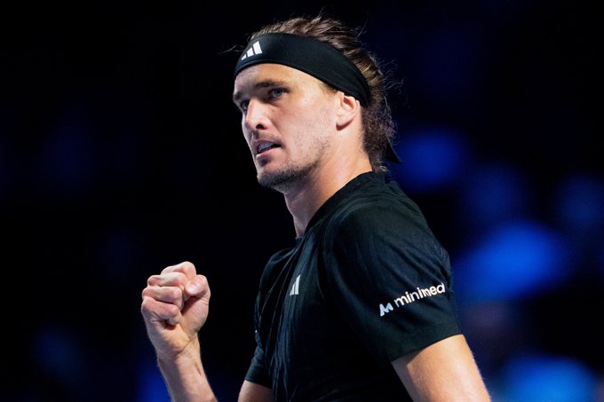 Alexander Zverev stands on tennis court wearing black Adidas shirt with Munich logo on sleeve, black headband over long hair, making fist pump gesture with right hand clenched, focused expression, dark background with blue lighting.
