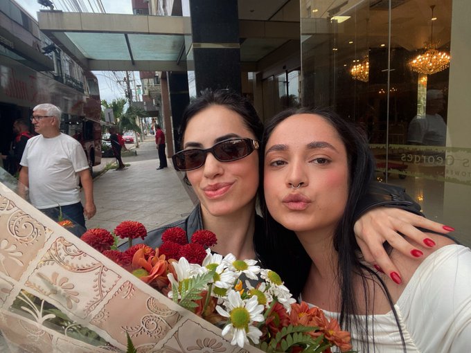 First image shows two women one older with dark hair wearing sunglasses and leather jacket the other younger with long dark hair in white top both smiling and posing closely outdoors on a sidewalk near a building entrance with red flowers and white daisies in a bouquet held by the younger woman elderly man in white shirt visible in background cars and urban architecture around. Second image captures two women embracing one with hair in bun wearing sunglasses and leather jacket the other with loose hair in white top holding a large bouquet of mixed flowers including red orange and white blooms wrapped in red paper against a red brick wall with another woman in pink pants nearby. Third image features two women standing side by side both with long dark hair one smiling widely in black top and leather jacket the other wearing sunglasses in black top holding a bouquet of red roses and white daisies wrapped in red paper against a red brick wall with ferns and greenery accents.