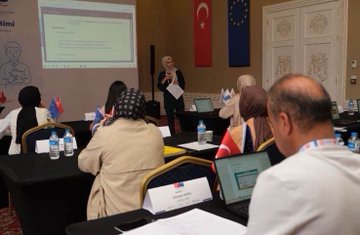 First image shows a large group of diverse participants including men and women in professional attire some wearing headscarves holding certificates seated and standing around black-draped tables with Turkish and EU flags in a grand room with chandeliers and wooden floors. Second image depicts participants engaged in a session with a male speaker in a suit gesturing at the front tables covered in documents water bottles and Turkish flags banners of UN and other organizations on walls in an ornate room with arched windows. Third image features a female speaker in headscarf holding notes in front of a projector screen displaying text on education with participants at tables using laptops water bottles and name cards Turkish flag and EU flag visible diverse group including headscarf-wearers. Fourth image shows a female presenter in headscarf holding a whiteboard drawing of cartoon figures a man and woman with books and a child pointing to educational text on screen Turkish EU and other flags below blue line drawings of professionals on wall.