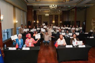 First image shows a large group of diverse participants including men and women in professional attire some wearing headscarves holding certificates seated and standing around black-draped tables with Turkish and EU flags in a grand room with chandeliers and wooden floors. Second image depicts participants engaged in a session with a male speaker in a suit gesturing at the front tables covered in documents water bottles and Turkish flags banners of UN and other organizations on walls in an ornate room with arched windows. Third image features a female speaker in headscarf holding notes in front of a projector screen displaying text on education with participants at tables using laptops water bottles and name cards Turkish flag and EU flag visible diverse group including headscarf-wearers. Fourth image shows a female presenter in headscarf holding a whiteboard drawing of cartoon figures a man and woman with books and a child pointing to educational text on screen Turkish EU and other flags below blue line drawings of professionals on wall.