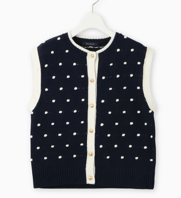 A navy sleeveless knit vest with white polka dots and gold buttons hangs on a white hanger against a white background. The vest has a crew neckline with white trim and button front closure. It appears to be the top worn by Yuko Araki in a TV show preview from the brand BRATRICE.