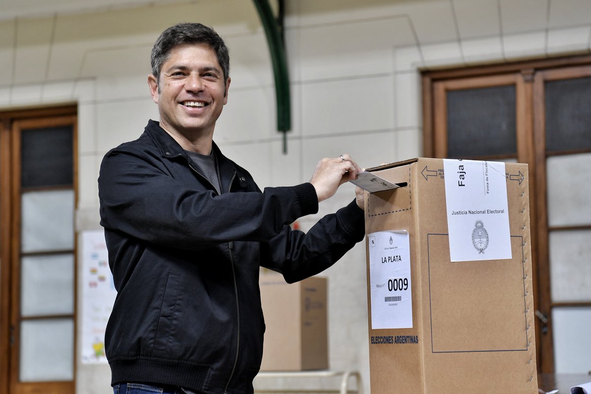 A man with short gray hair wearing a dark jacket stands in a room with white tiled walls and wooden doors holding a ballot slip toward a cardboard ballot box labeled with official election papers including Ficha 003 and La Plata on the side with a green archway in the background.