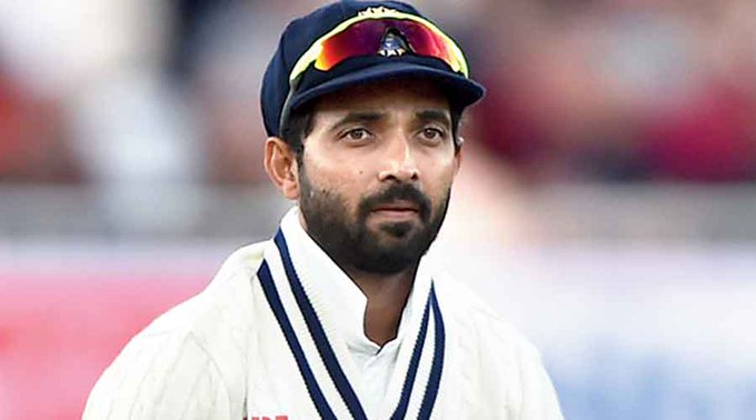 Ajinkya Rahane stands on a cricket field wearing a white uniform with blue accents a navy blue cap with yellow emblem and yellow-tinted sunglasses looking focused with a blurred stadium crowd in the background.