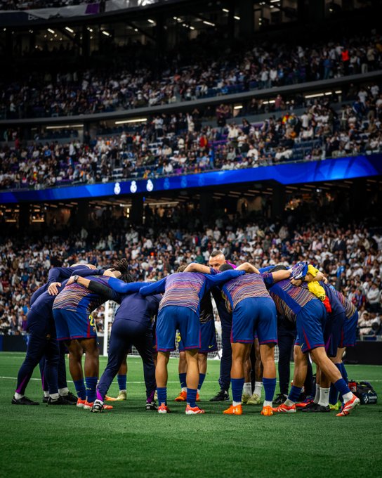 Group of soccer players in blue and red striped jerseys and shorts stand in a tight huddle on green field embracing each other shoulder to shoulder facing away from camera with arms linked coach in dark suit nearby stadium filled with spectators in background blue lighting on upper stands goal net visible behind them orange and white cleats on feet