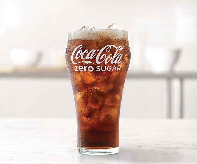 A tall glass filled with dark cola liquid, ice cubes, and foam, featuring the Coca-Cola Zero Sugar logo on the glass; placed on a white table in a bright, minimalist kitchen setting with blurred background elements like cups and a counter.