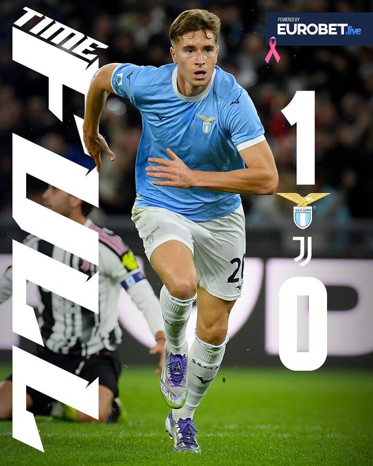 A male soccer player with short blond hair wearing a light blue Lazio jersey numbered 20, white shorts, purple socks, and cleats runs on a green field during a match against Juventus. He has a focused expression and is in motion with one leg forward. In the background, blurred Juventus players in black and white stripes are visible on the field. Overlaid graphics include the score 1-0 for Lazio, the match name Lazio Juventus, a pink ribbon, and sponsor Eurobet live branding.