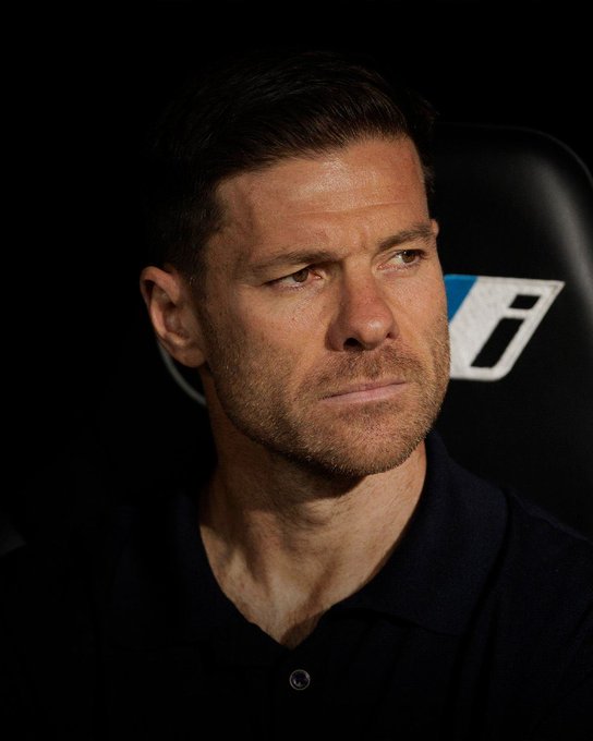 A man with short dark hair and a serious expression sits in a black chair against a dark background wearing a dark blue polo shirt with a small white logo on the chest. The setting appears to be an indoor sports or press area with subtle lighting highlighting his face.