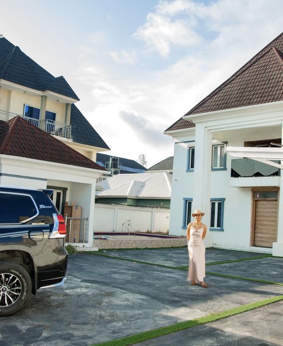 A woman in a light-colored dress and wide-brim hat stands on a paved driveway in front of a large white multi-story house with blue windows, beige accents, and a wooden door. Adjacent is a beige building with red roof tiles and balconies. A dark blue SUV is parked nearby with its door open. The scene is outdoors under a partly cloudy sky with green grass patches.