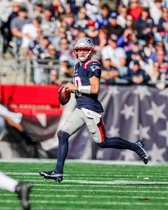 Player wearing navy blue New England Patriots jersey number 10 and helmet runs with football on green football field during game with crowd in blue and red stands in background and American flag visible.