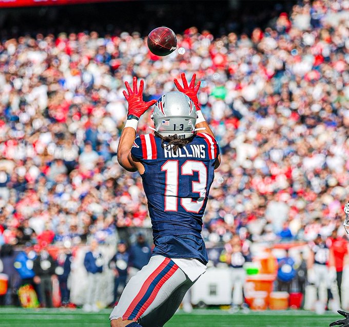 A football player wearing a navy blue New England Patriots jersey with number 13 and name Collins on the back stands on a green field during a game with a large crowd in the stadium background he extends red gloves upward to catch a brown football above his silver helmet