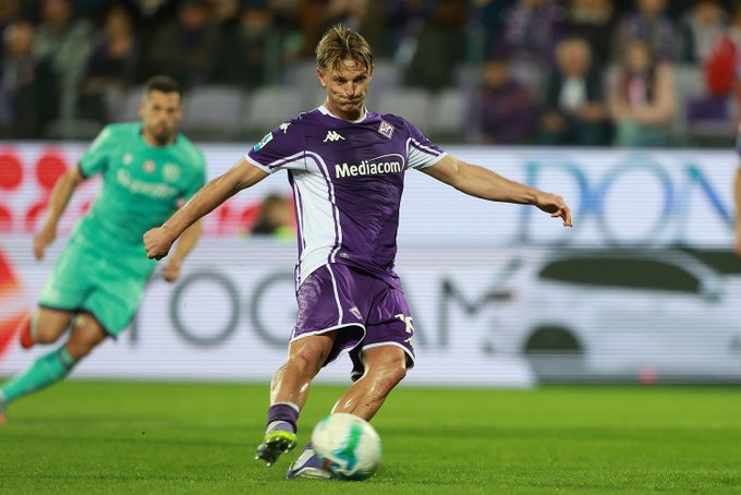 A soccer player in a purple and white Fiorentina jersey with Mediocom sponsor kicks a green and white ball during a match on a green field while another player in a green Bologna jersey stands nearby with a blurred stadium crowd and advertisements in the background.