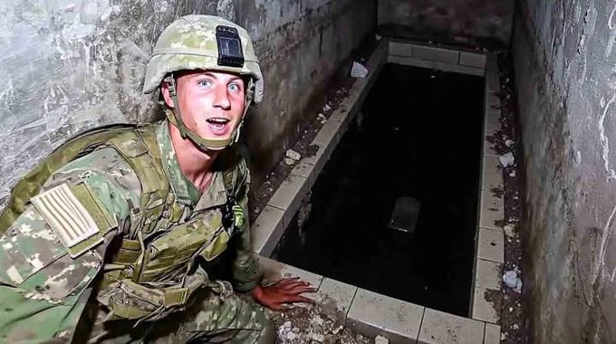 The image shows a U.S. soldier in multicam camouflage uniform, helmet with night vision mount, and tactical gear, crouching and peering intently into a dark rectangular opening (possibly a manhole or tunnel entrance) in a concrete floor within a dimly lit, graffiti-covered underground or basement area with tiled walls and debris.
