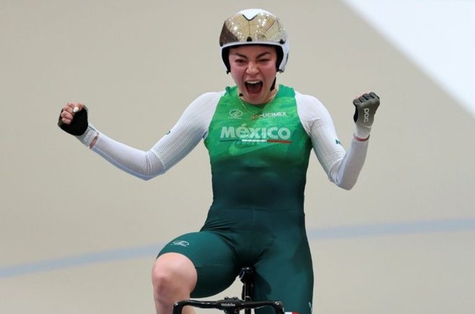 Female cyclist in green Mexico branded uniform and shorts sits on bike with arms raised in victory yell, wearing white helmet with gold accents and gloves, on indoor track with white floor and blurred background.