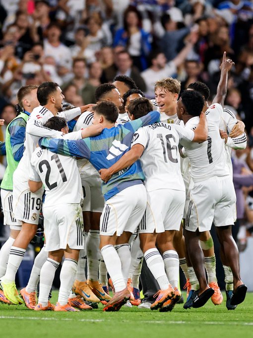 Group of soccer players in white and blue jerseys including numbers 21, 19, 18, and 11 huddle together on green field raising arms in celebration surrounded by stadium crowd in background.