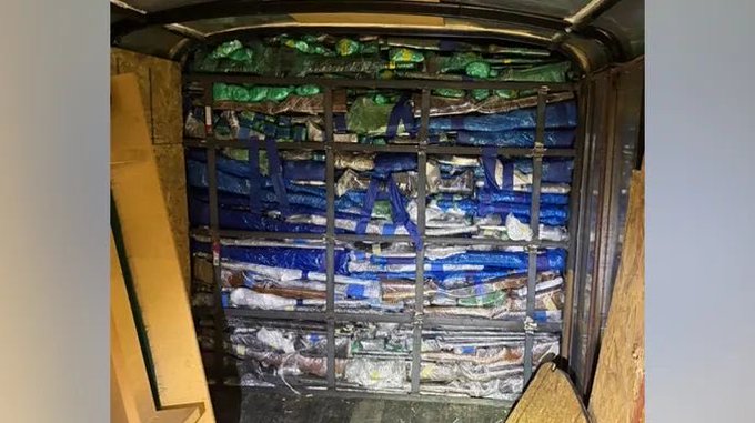 First image shows numerous firearms of various types including rifles shotguns and handguns laid out on tables covered with blue plastic bags in an indoor warehouse setting with law enforcement officers in uniforms examining and handling them surrounded by cardboard boxes and tables. Second image depicts the interior of a wooden trailer with walls lined by multiple blue plastic-wrapped bundles secured with straps indicating hidden compartments used for smuggling the weapons.