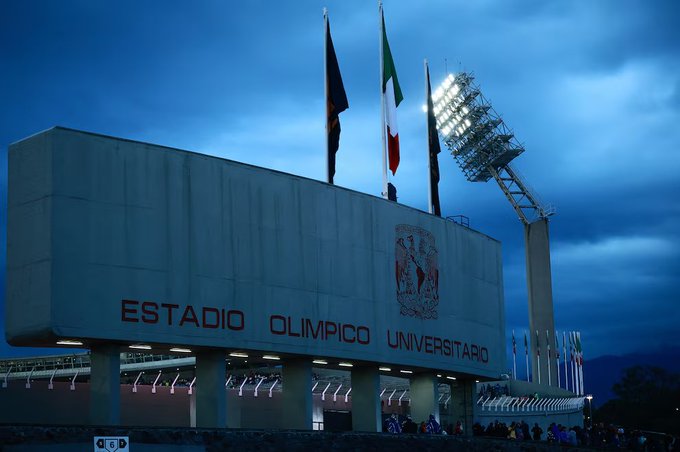 Large white stadium building with Estadio Olímpico Universitario text and university emblem on the facade, multiple international flags including Italian, French, and Mexican on poles, bright floodlights illuminating the structure, people visible in the foreground under a dark cloudy sky at night.