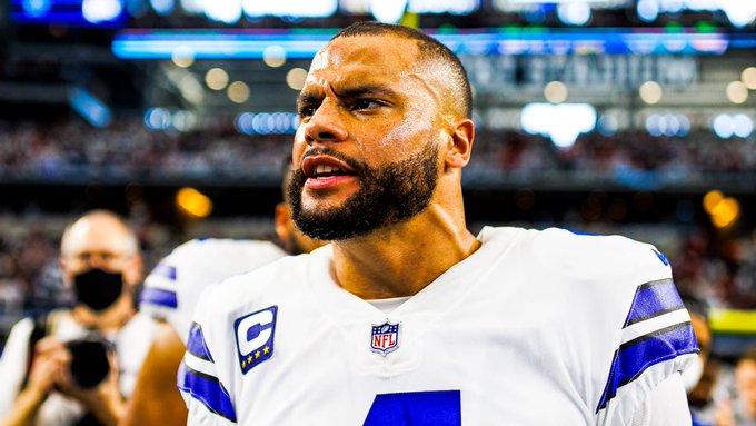 A man with short hair and a beard wearing a white Dallas Cowboys NFL jersey with number 1 and captain C patch stands on a football field during a game with stadium lights and crowd in the blurred background another person in a mask is nearby