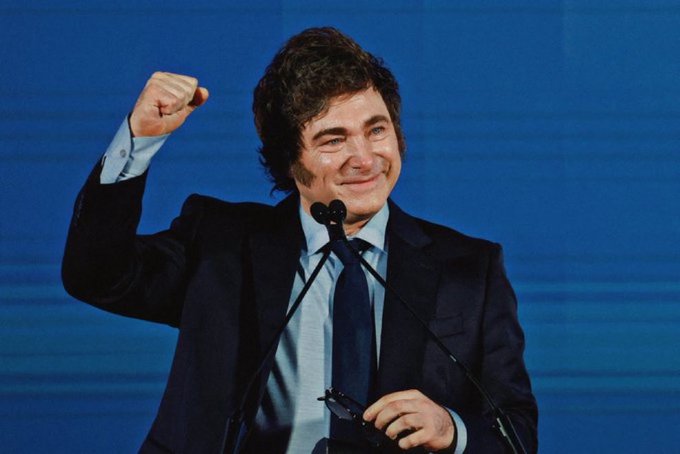 A man with curly dark hair wearing a dark suit white shirt and blue tie stands at a podium with a microphone raising his right fist in a celebratory gesture while smiling his left hand holds a microphone against a solid blue background