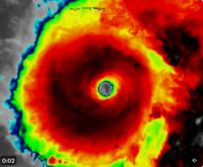 Satellite image displays Hurricane Melissa as a large swirling storm system with a distinct eye at the center surrounded by intense red and yellow bands indicating heavy cloud cover and strong winds extending outward in a circular pattern over the ocean south of Jamaica with color-coded intensity gradients from gray outer edges to vibrant inner core