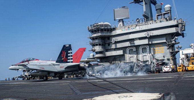 A US Navy aircraft carrier deck on the USS Nimitz with a gray F/A-18E/F Super Hornet fighter jet positioned near the edge showing red white and blue tail markings. Smoke and flames rise from the area behind the jet indicating a crash or fire incident. An MH-60 Seahawk helicopter is visible nearby with its rotors and fuselage intact. The carrier structure includes multiple levels railings antennas and equipment in the background under a clear blue sky.