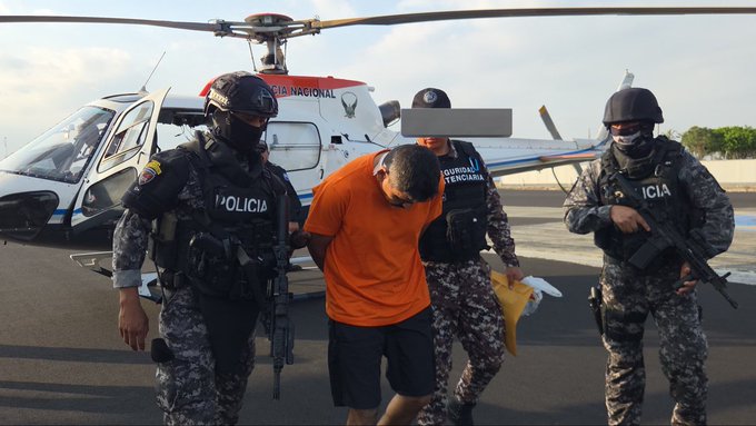 Several police officers in black tactical vests and helmets, some wearing masks, escort a man in an orange shirt and shorts handcuffed behind his back near a white police helicopter on a tarmac. One officer carries a rifle, another holds a bag. The man appears to be under arrest, with officers positioned around him. The helicopter has Policia markings on its side.