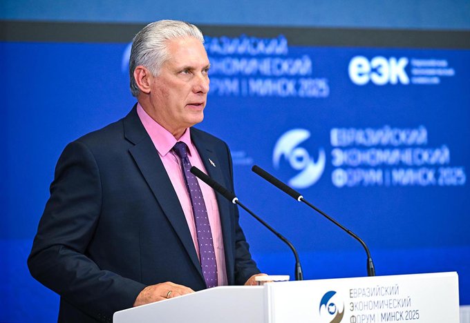 A man with gray hair wearing a dark suit and pink tie stands at a podium speaking into microphones, with a blue backdrop displaying text in Russian including E3K and dates like МАРТ 2025, and logos of organizations.