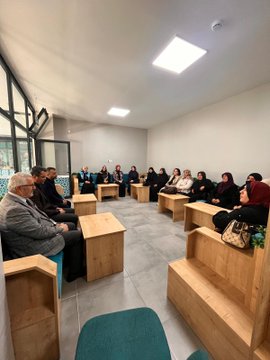 First image shows a group of men and women seated in a circle on wooden benches in a modern room with large windows and fluorescent lighting, engaged in discussion, some wearing headscarves and formal clothing. Second image depicts women in black attire and headscarves sitting around wooden tables with plates of food, suggesting a meal or tea session. Third image captures similar group discussion setup with participants including elderly men in suits and women in modest dress. Fourth image features a group of women in headscarves and vests standing together in front of wooden bookshelves with plants and blue boxes on a table, posed for a photo.