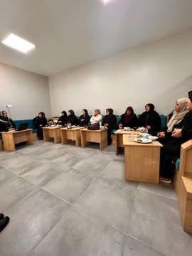 First image shows a group of men and women seated in a circle on wooden benches in a modern room with large windows and fluorescent lighting, engaged in discussion, some wearing headscarves and formal clothing. Second image depicts women in black attire and headscarves sitting around wooden tables with plates of food, suggesting a meal or tea session. Third image captures similar group discussion setup with participants including elderly men in suits and women in modest dress. Fourth image features a group of women in headscarves and vests standing together in front of wooden bookshelves with plants and blue boxes on a table, posed for a photo.