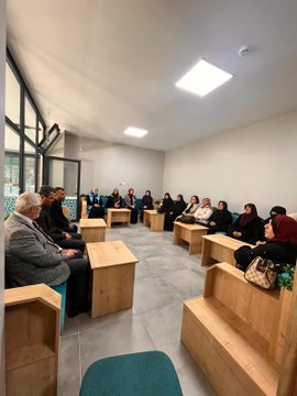 First image shows a group of men and women seated in a circle on wooden benches in a modern room with large windows and fluorescent lighting, engaged in discussion, some wearing headscarves and formal clothing. Second image depicts women in black attire and headscarves sitting around wooden tables with plates of food, suggesting a meal or tea session. Third image captures similar group discussion setup with participants including elderly men in suits and women in modest dress. Fourth image features a group of women in headscarves and vests standing together in front of wooden bookshelves with plants and blue boxes on a table, posed for a photo.