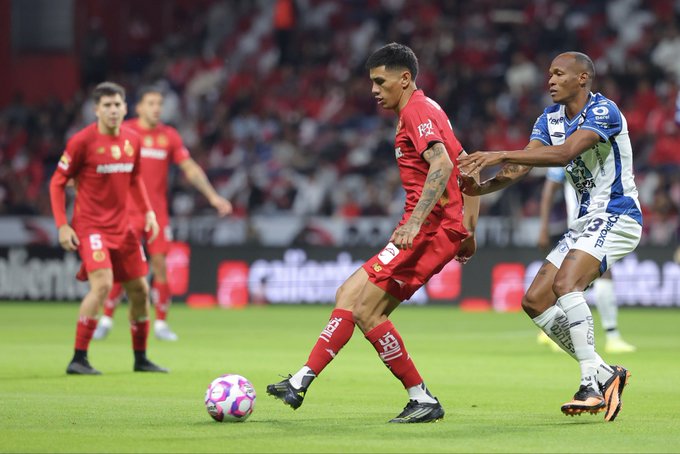 Soccer players in red jerseys from Toluca compete against those in blue and white from Pachuca on a green field at a stadium filled with spectators. A player in red number 6 kicks a purple and white ball while another in blue grabs his arm. Additional players in red stand nearby, and the scene captures dynamic action during the match.