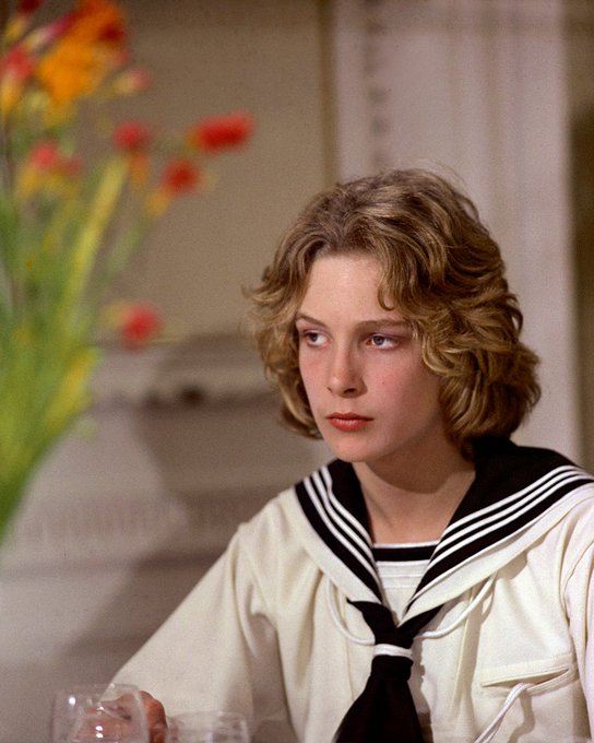First image shows a young boy with curly blonde hair wearing a white sailor uniform with black collar sitting at a table holding a glass surrounded by red and orange flowers in a room with ornate walls. Second image depicts the same boy in a dark naval uniform with gold buttons standing in a dimly lit area with a spotlight nearby. Third image features the boy in a black military-style jacket with buttons viewed through window bars in a dark setting.