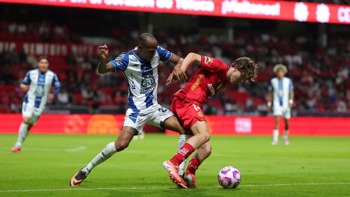 Two soccer players in action on a green field during a match, one in blue and white striped jersey of Toluca tackling another in red jersey of Pachuca near a purple and white ball, with stadium crowd and scoreboard in background.