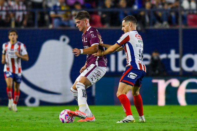Two soccer players in action on a green field during a match one in maroon jersey with white pants marked Necaxa grabs the arm of the other in white and blue striped jersey marked Atlético San Luis while reaching for a pink and white soccer ball near the goal line with other players and stadium seating in the background.
