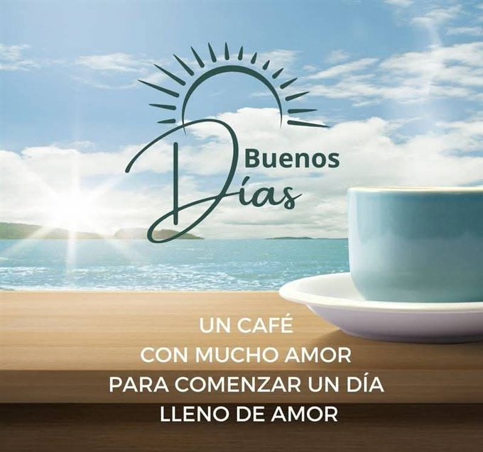 A serene outdoor scene with a calm sea under a partly cloudy blue sky and a bright sun low on the horizon. In the foreground on a wooden table sits a light blue ceramic coffee cup on a matching saucer containing a hot beverage with steam rising. Elegant black cursive text overlays read Buenos dias at the top near the sun, un cafe con mucho amor para comenzar un dia lleno de amor below the cup.