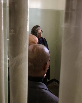 First image shows a woman in a beige trench coat and jeans standing in front of a framed display with two black and white photographs on a beige wall outdoors. The left photo depicts a line of people in uniforms walking in a row. The right photo shows a woman in military attire standing next to a man in a suit. Second image depicts an older woman with gray hair wearing glasses and a dark outfit standing next to a bald man in a dark suit inside a room with green walls and metal bars in the background. Third image shows four people standing on steps outside a gray door with a yellow sign two men and two women dressed in casual and formal attire including jackets and a shirt with African motifs.