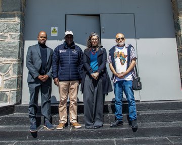 First image shows a woman in a beige trench coat and jeans standing in front of a framed display with two black and white photographs on a beige wall outdoors. The left photo depicts a line of people in uniforms walking in a row. The right photo shows a woman in military attire standing next to a man in a suit. Second image depicts an older woman with gray hair wearing glasses and a dark outfit standing next to a bald man in a dark suit inside a room with green walls and metal bars in the background. Third image shows four people standing on steps outside a gray door with a yellow sign two men and two women dressed in casual and formal attire including jackets and a shirt with African motifs.