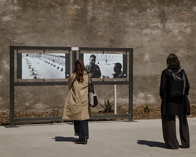 First image shows a woman in a beige trench coat and jeans standing in front of a framed display with two black and white photographs on a beige wall outdoors. The left photo depicts a line of people in uniforms walking in a row. The right photo shows a woman in military attire standing next to a man in a suit. Second image depicts an older woman with gray hair wearing glasses and a dark outfit standing next to a bald man in a dark suit inside a room with green walls and metal bars in the background. Third image shows four people standing on steps outside a gray door with a yellow sign two men and two women dressed in casual and formal attire including jackets and a shirt with African motifs.