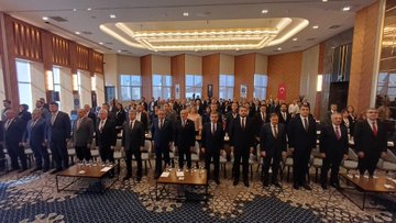 First image shows a group of men in suits standing on a stage with a backdrop featuring Turkish flags, blue and red panels, and a logo for an event. They pose formally in front of a wooden-paneled wall. Second image depicts a man in a suit speaking at a podium holding a microphone, with a large screen behind showing text about Gün Sazak merit and honesty essay contest in public service. The setting includes wooden walls and a blue screen. Third image displays a large group of people, mostly men in suits, standing in rows in a grand hall with ornate ceilings and wooden floors, some wearing badges. Fourth image features two men in suits on stage, one presenting a red box with a plaque to the other, against a blue backdrop with text about Gün Sazak award and 1992, including a logo.