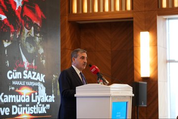 First image shows a group of men in suits standing on a stage with a backdrop featuring Turkish flags, blue and red panels, and a logo for an event. They pose formally in front of a wooden-paneled wall. Second image depicts a man in a suit speaking at a podium holding a microphone, with a large screen behind showing text about Gün Sazak merit and honesty essay contest in public service. The setting includes wooden walls and a blue screen. Third image displays a large group of people, mostly men in suits, standing in rows in a grand hall with ornate ceilings and wooden floors, some wearing badges. Fourth image features two men in suits on stage, one presenting a red box with a plaque to the other, against a blue backdrop with text about Gün Sazak award and 1992, including a logo.