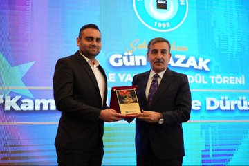 First image shows a group of men in suits standing on a stage with a backdrop featuring Turkish flags, blue and red panels, and a logo for an event. They pose formally in front of a wooden-paneled wall. Second image depicts a man in a suit speaking at a podium holding a microphone, with a large screen behind showing text about Gün Sazak merit and honesty essay contest in public service. The setting includes wooden walls and a blue screen. Third image displays a large group of people, mostly men in suits, standing in rows in a grand hall with ornate ceilings and wooden floors, some wearing badges. Fourth image features two men in suits on stage, one presenting a red box with a plaque to the other, against a blue backdrop with text about Gün Sazak award and 1992, including a logo.