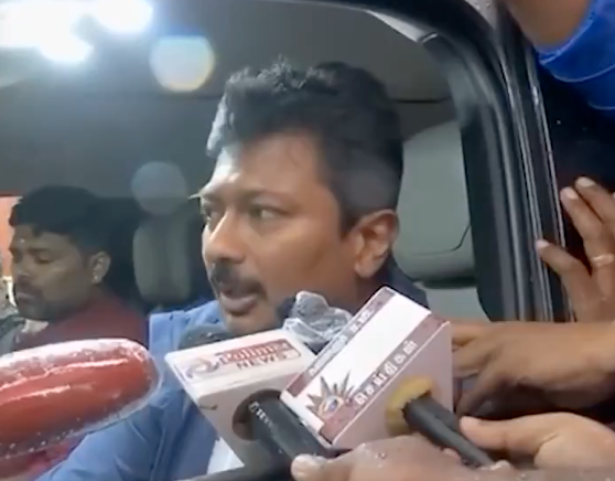 A man in a blue shirt and glasses speaks into a microphone labeled News 18 inside a car with another man visible nearby holding a microphone. The setting includes a window showing exterior light and another person outside. The microphone has Tamil text on it.
