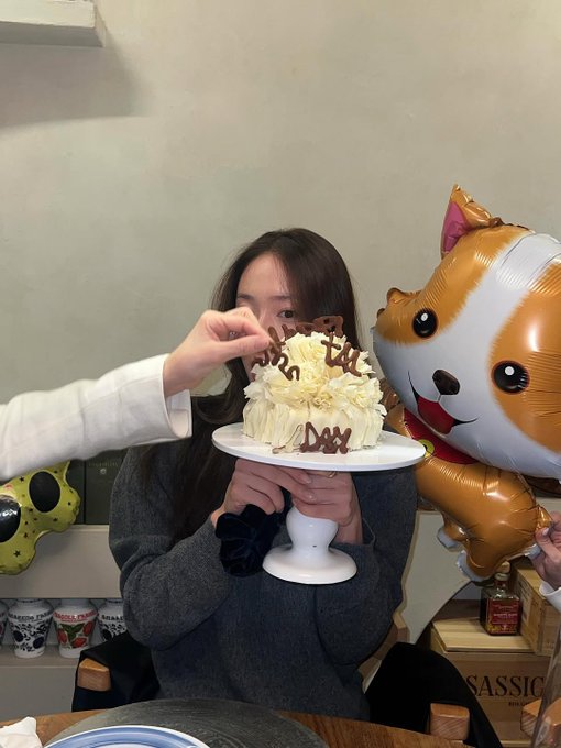 In the first image a white cake topped with chocolate writing sits on a stand beside a large orange and white balloon shaped like a dog with floppy ears and a smiling face while a hand blows out sparklers on top and another hand holds a streamer nearby with beige walls and wooden elements in the background. In the second image two young women with long dark hair wear casual gray sweaters and take a close-up selfie smiling with one wearing a black beanie with a logo while holding a phone the other makes a playful expression next to a white cake with chocolate writing on a glass table with blue plates around. In the fourth image a young woman with long dark hair in a black blazer holds a beige card with handwritten text close to her face smiling slightly with focused eyes while sitting near a white gift box and gold swirled balloons against a beige wall with shelves.