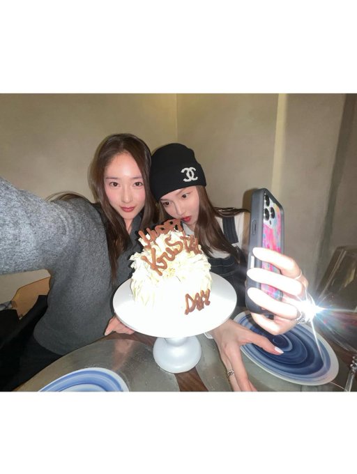 In the first image a white cake topped with chocolate writing sits on a stand beside a large orange and white balloon shaped like a dog with floppy ears and a smiling face while a hand blows out sparklers on top and another hand holds a streamer nearby with beige walls and wooden elements in the background. In the second image two young women with long dark hair wear casual gray sweaters and take a close-up selfie smiling with one wearing a black beanie with a logo while holding a phone the other makes a playful expression next to a white cake with chocolate writing on a glass table with blue plates around. In the fourth image a young woman with long dark hair in a black blazer holds a beige card with handwritten text close to her face smiling slightly with focused eyes while sitting near a white gift box and gold swirled balloons against a beige wall with shelves.