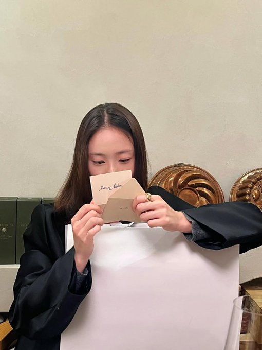 In the first image a white cake topped with chocolate writing sits on a stand beside a large orange and white balloon shaped like a dog with floppy ears and a smiling face while a hand blows out sparklers on top and another hand holds a streamer nearby with beige walls and wooden elements in the background. In the second image two young women with long dark hair wear casual gray sweaters and take a close-up selfie smiling with one wearing a black beanie with a logo while holding a phone the other makes a playful expression next to a white cake with chocolate writing on a glass table with blue plates around. In the fourth image a young woman with long dark hair in a black blazer holds a beige card with handwritten text close to her face smiling slightly with focused eyes while sitting near a white gift box and gold swirled balloons against a beige wall with shelves.
