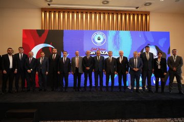 First image shows a man in a dark suit speaking at a podium with a microphone, gesturing with his hand raised, against a blue backdrop featuring the text Gün Sazak Makale Yarışması Ödül Töreni and a 1992 emblem. Second image depicts a group of about 15 people in formal suits and one woman in professional attire standing together on a stage with Turkish flags, red and blue banners, and a logo possibly for MHP. Third image captures a bald man in glasses and a dark suit holding a microphone at a white podium, gesturing openly, with a large projected image of another man behind him and wooden paneling in the background. Fourth image features two men in suits, one with a mustache receiving a red plaque award from the other, standing before a purple and blue backdrop with Turkish flags and text for Sabiha Gökçen Gün Sazak Liyakat ve Dürüstlük Makale Yarışması.