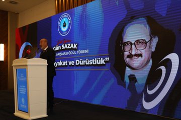 First image shows a man in a suit speaking at a podium with a microphone in front of a blue backdrop featuring the Gün Sazak name and themes of merit and honesty in public service, alongside a large portrait of Gün Sazak wearing glasses and a mustache. Second image depicts a man in a suit holding a microphone at a podium with a wooden backdrop and a large black-and-white portrait of Gün Sazak, text reading Gün Sazak Makale Yarışması and Kamuda Liyakat ve Dürüstlük. Third image displays a man in a suit speaking at a podium with a microphone, blue backdrop with Türk Büro-Sen logo and Gün Sazak portrait, text on merit and honesty in public service. Fourth image features a group of about 15 people in formal suits and dresses standing on a stage with a blue backdrop displaying a circular emblem.